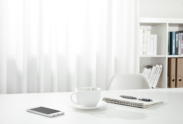 Primer plano de una taza de café, un cuaderno y un bolígrafo y un teléfono inteligente en un escritorio blanco, en el interior