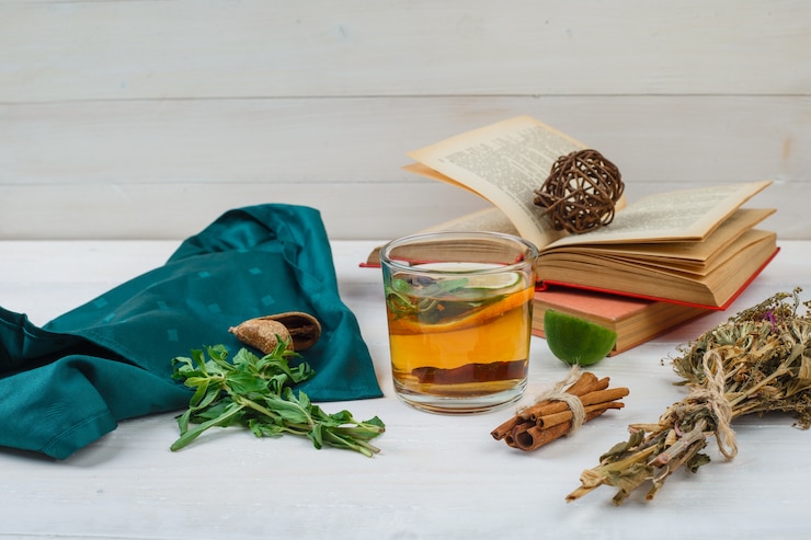 Primer plano de té de hierbas y flores con libros, limón, especias y bufanda verde