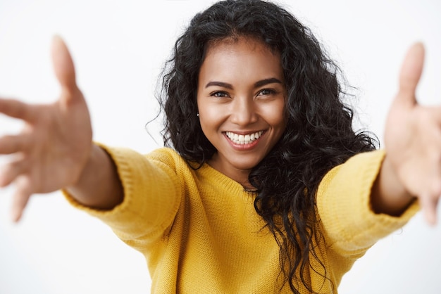 Primer plano tierna y linda mujer afroamericana de pelo rizado con suéter amarillo estirando las manos hacia adelante sonriendo y mirando la cámara con amor y cuidado para dar un abrazo a un amigo