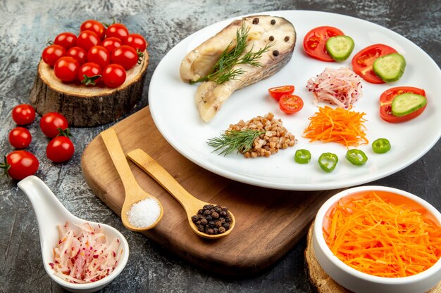 Primer plano de trigo sarraceno de pescado hervido servido con verduras verdes en un plato blanco sobre una tabla de cortar de madera y especias en la superficie del hielo