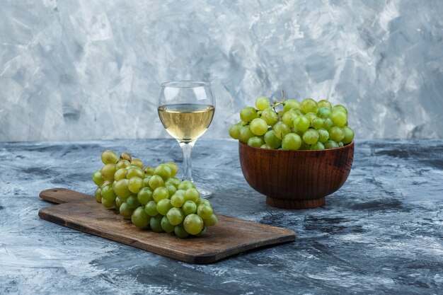 Primer plano de uvas blancas en un tazón con copa de vino, uvas en una tabla de cortar sobre fondo de mármol azul claro y oscuro. horizontal
