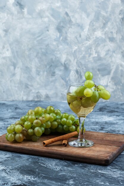 Primer plano de uvas blancas, vaso de whisky, canela en tabla de cortar sobre fondo de mármol azul claro y oscuro. vertical