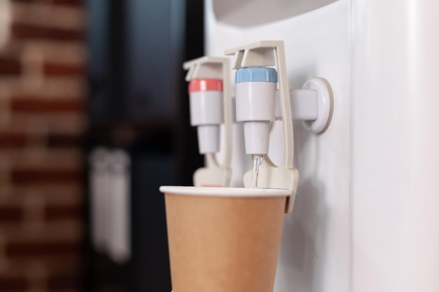 Foto gratuita primer plano de un vaso desechable en el enfriador de agua en la oficina de inicio. dispensador de agua con grifo frío y caliente utilizado para refrescos y bebidas en el espacio. agua mineral purificada en galón.