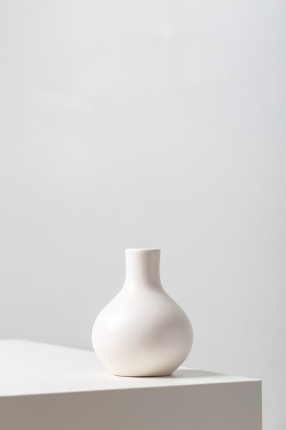 Primer plano vertical de una vasija de arcilla blanca sobre la mesa bajo las luces sobre un fondo blanco.
