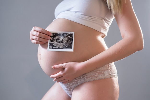 Primer plano del vientre del modelo embarazado joven que muestra la imagen ultrasónica de su bebé. Futura mamá en su segundo trimestre que sostiene la exploración del ultrasonido de su niño. Concepto de maternidad