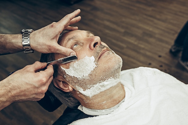 Primer plano vista superior lateral guapo barbudo senior hombre caucásico barba preparación en la moderna barbería. Peluquería al servicio del cliente, corte de pelo de barba con navaja de afeitar. Concepto de peluquería.