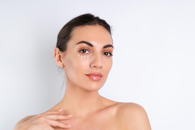 Primer retrato de belleza de una mujer en topless con piel perfecta y maquillaje natural con puntos de crema antienvejecimiento para hidratar y reafirmar la piel debajo de los ojosx9