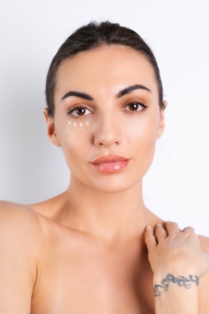 Primer retrato de belleza de una mujer en topless con piel perfecta y maquillaje natural con puntos de crema antienvejecimiento para hidratar y reafirmar la piel debajo de los ojosx9