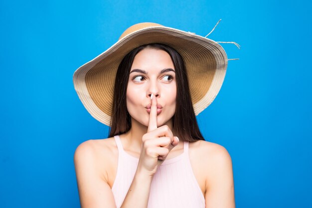 Foto gratuita primer retrato de mujer con sombrero de paja mostrando el dedo en los labios para mantener el secreto aislado sobre la pared azul.