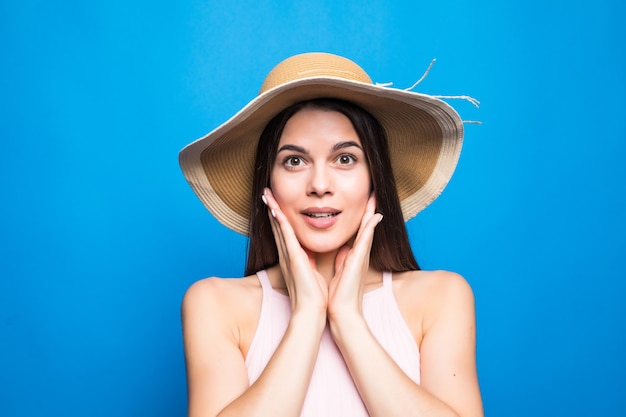 Primer retrato de mujer sorprendida con sombrero de paja con las manos en las mejillas aisladas sobre la pared azul.