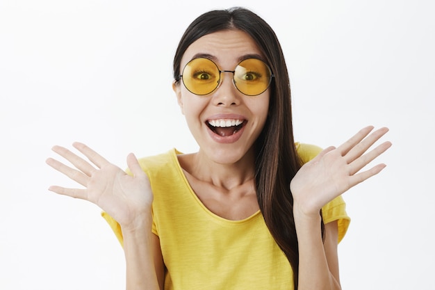 Primer tiro de encantadora mujer elegante encantada en gafas de sol de moda y camiseta amarilla agitando las palmas levantadas cerca de los hombros