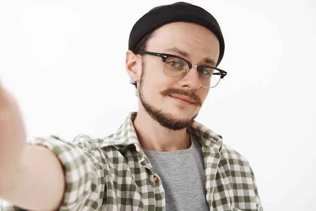 Primer tiro de hombre guapo joven coqueto seguro de sí mismo con barba en gorro negro elegante y camisa a cuadros tomando selfie