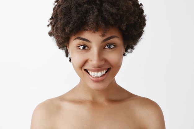 Primer tiro de mujer afroamericana confiada y femenina coqueta con amplia sonrisa brillante posando desnuda satisfecha con gran condición de la piel después del tratamiento cosmetológico