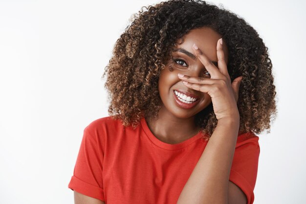 Primer tiro de mujer morena tierna y femenina divertida con corte de pelo rizado cerca de la mitad de la cara con la mano y sonriendo, riendo disfrutando de una gran compañía divertida sobre fondo blanco. Copia espacio