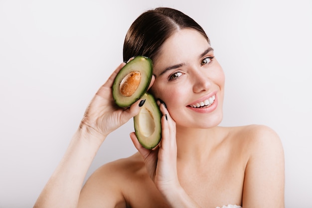 Primer tiro de mujer sonriente de ojos verdes sin maquillaje en la pared blanca. Modelo demuestra los beneficios de la piel del aguacate.