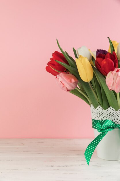 Primer de tulipanes en el florero blanco con el arco verde en el escritorio de madera contra fondo rosado