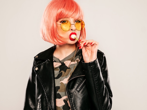 Primeros labios de la joven y bella chica mala inconformista en una chaqueta de cuero negro de moda y un arete en la nariz. Mujer sonriente despreocupada y sexy posando en el estudio con peluca rosa. Modelo positivo lamiendo caramelos redondos