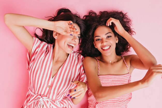 Los primos de muy buen humor yacen en el suelo de buen humor. Las niñas posan con sonrisas sinceras en ropa rosa para retratos de primer plano.