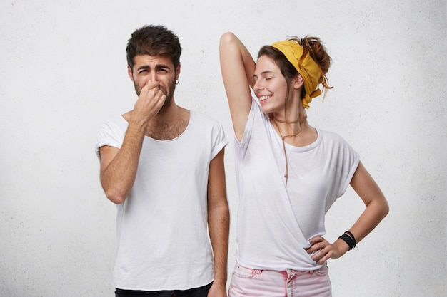 Foto gratuita problemas con el olor corporal. hombre disgustado pellizcando su nariz sintiendo mal olor o hedor que sale de una atractiva chica sonriente, que está levantando su brazo, mostrando una camiseta mojada debido al sudor de las axilas