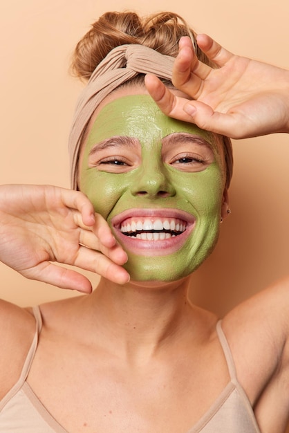 Procedimientos de belleza y concepto de cuidado de la piel. Mujer alegre mantiene las manos en la cara sonríe positivamente muestra dientes blancos disfruta de tratamiento facial aplica máscara nutritiva verde usa poses de diadema interior