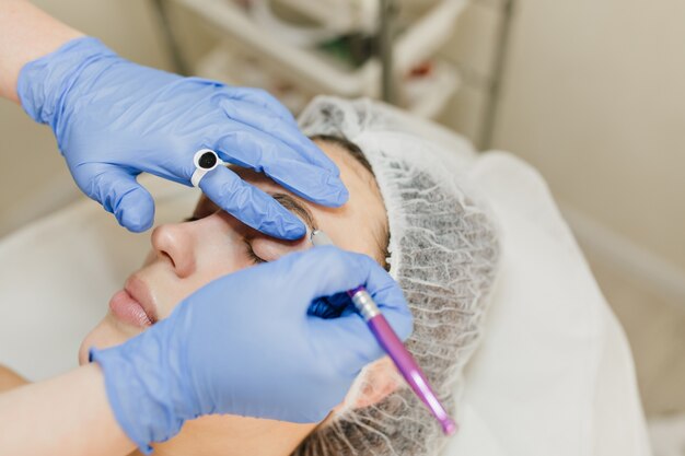 Procedimientos de cosmetología de mujer joven y bonita en salón de belleza. Procedimiento de dermatología, pintura de cejas, manos en resplandor azul, en el trabajo, cuidado de la salud, terapia, botox, belleza