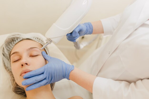 Procedimientos de cosmetología, rejuvenecimiento de mujer joven y bonita en salón de belleza. Procedimiento de dermatología, manos en resplandor azul, en el trabajo, atención médica, terapia, botox, inyecciones