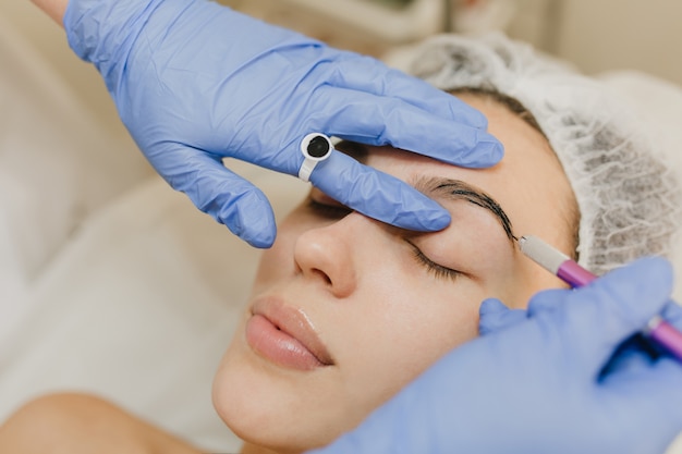 Procedimientos de cosmetología de retrato de mujer joven y bonita en salón de belleza. Procedimiento de dermatología, cejas de botox, manos en resplandores azules, en el trabajo, cuidado de la salud, terapia, belleza, rejuvenecimiento