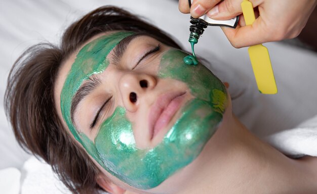 El proceso de aplicar una mascarilla cosmética verde en el rostro de una mujer joven, procedimiento de spa en el salón, belleza y cuidado de la piel.