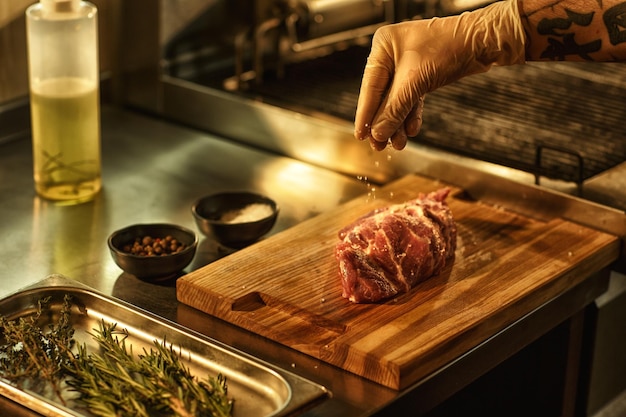 Proceso de preparación de bistec de ternera en un chef profesional de cocina de restaurante con guante blanco que sala la carne con ingredientes como condimentos, aceite de pimienta y romero para un sabor picante