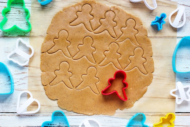 Proceso de tratar con las galletas de hombre de pan de jengibre, use molde de hombre de pan de jengibre rojo para cortar masa de pan de jengibre sobre papel de hornear alrededor de coloridos cortadores de galletas en la mesa de madera blanca. vista superior