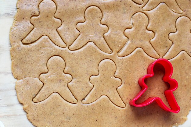 Proceso de tratar con las galletas de hombre de pan de jengibre, use molde de hombre de pan de jengibre rojo para cortar masa de pan de jengibre sobre papel de hornear alrededor de coloridos cortadores de galletas en la mesa de madera blanca. vista superior