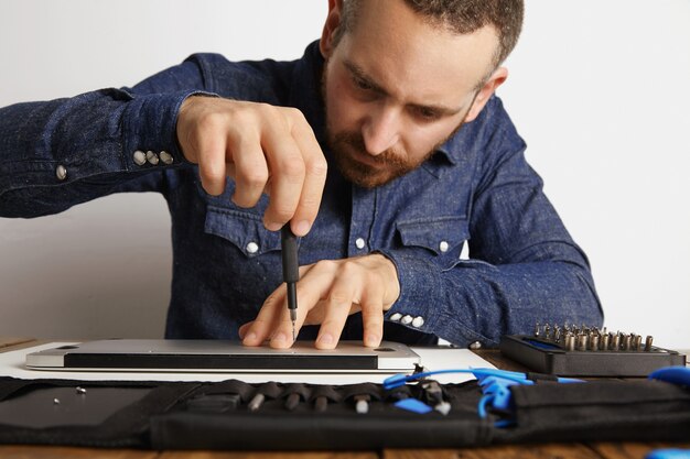 Profesional desenroscando con precisión la carcasa de la computadora portátil delgada metálica en su laboratorio de servicio eléctrico cerca de la bolsa de herramientas para limpiarla y repararla, vista frontal