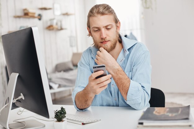 El profesional independiente masculino de barba rubia instala una nueva aplicación en el teléfono inteligente, descarga el programa en la computadora, usa wi-fi y recibe el mensaje del socio. Negocios, tecnologías modernas, comunicación.