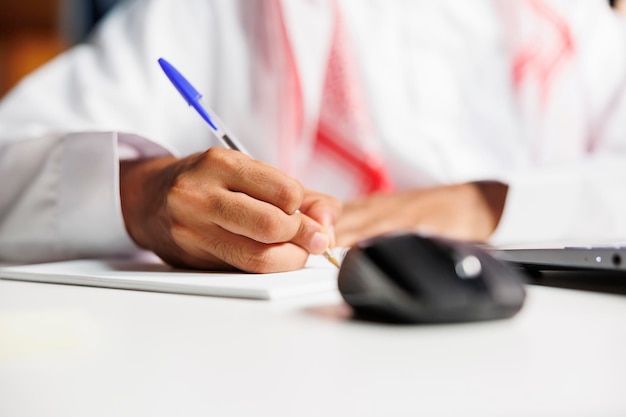 Foto gratuita un profesional independiente de oriente medio trabaja eficientemente en su escritorio, garabateando en su cuaderno. realiza investigaciones en línea, toma notas y se mantiene conectado mediante tecnología inalámbrica.