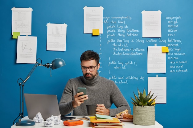 El profesional de TI trabaja en un proyecto de inicio, actualiza el software y la base de datos en el teléfono móvil, bebe bebidas calientes, se sienta en el escritorio contra la pared azul con información escrita.