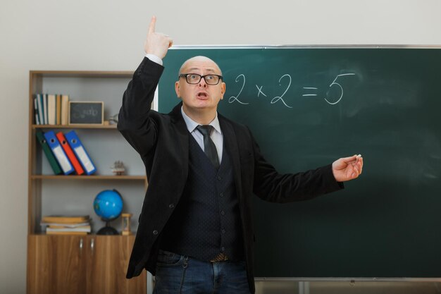 Profesor con anteojos parado cerca de la pizarra en el aula explicando la lección emocionado señalando con el dedo índice hacia arriba con una nueva idea