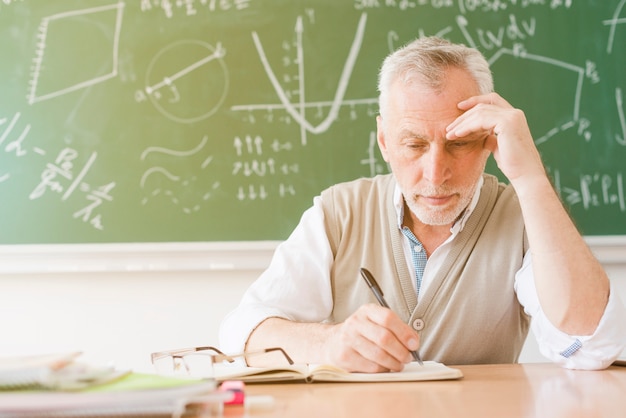 Profesor cansado envejecido que escribe en cuaderno en sala de clase