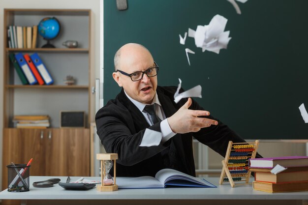 Profesor enojado con gafas sentado en el escritorio de la escuela frente a la pizarra en el aula arrojando trozos de papel