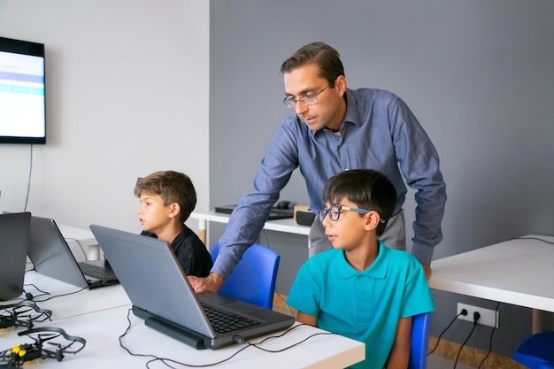 Foto gratuita profesor en gafas de pie detrás de la pupila y comprobación de tareas en portátil