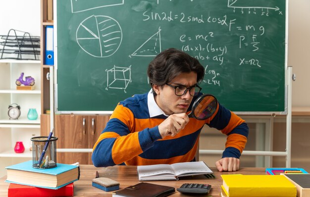 Profesor de geometría caucásico joven concentrado con gafas sentado en un escritorio con herramientas escolares en el aula manteniendo la mano en el escritorio mirando a un lado a través de una lupa