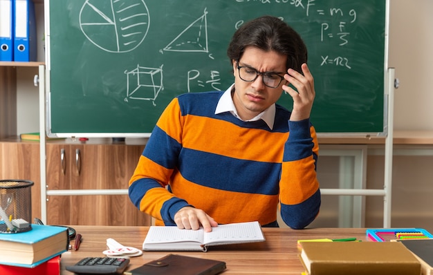 Profesor de geometría caucásico joven confundido con gafas sentado en el escritorio con herramientas escolares en el aula tocando gafas con el dedo señalador en el libro abierto leyéndolo