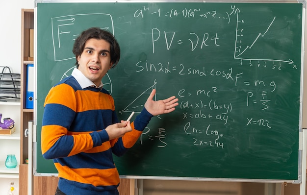 Profesor de geometría joven emocionado de pie en la vista de perfil delante de la pizarra en el aula sosteniendo tiza apuntando a la pizarra