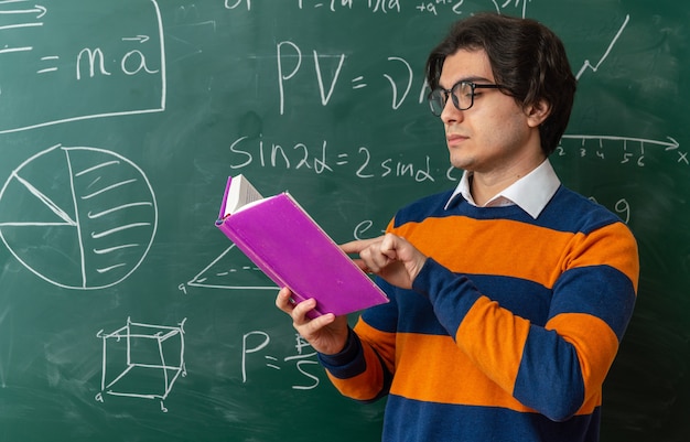 Profesor de geometría joven serio con gafas de pie delante de la pizarra en el aula libro de lectura apuntando con el dedo sobre él