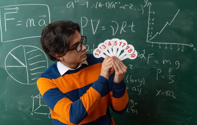 Profesor de geometría joven serio con gafas de pie delante de la pizarra en el aula sosteniendo y mirando los ventiladores de números