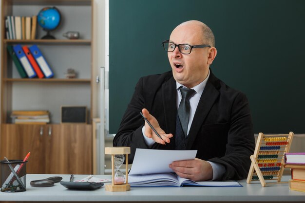 Profesor hombre con anteojos sentado en el escritorio de la escuela frente a la pizarra en el aula revisando la tarea de los estudiantes que se ven asombrados y conmocionados