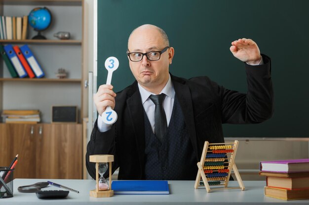 Profesor hombre con anteojos sentado en el escritorio de la escuela con registro de clase frente a la pizarra en el aula sosteniendo placas de matrícula explicando la lección confundida