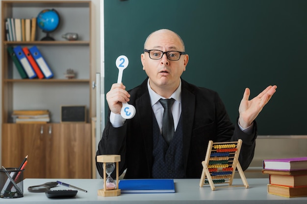 Profesor hombre con anteojos sentado en el escritorio de la escuela con registro de clase frente a la pizarra en el aula sosteniendo placas de matrícula explicando la lección que parece confundido con dudas