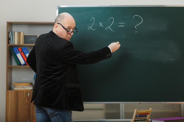 Profesor hombre con gafas de pie cerca de la pizarra en el aula explicando la lección confundido