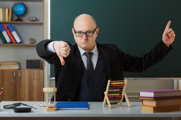 Profesor hombre con gafas con registro de clase sentado en el escritorio de la escuela frente a la pizarra en el aula con aspecto disgustado mostrando el pulgar hacia abajo apuntando con el dedo índice hacia arriba