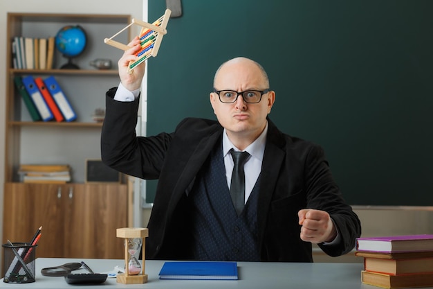 Profesor hombre con gafas con registro de clase sentado en el escritorio de la escuela frente a la pizarra en el aula balanceando el puño de ábaco enojado y frustrado